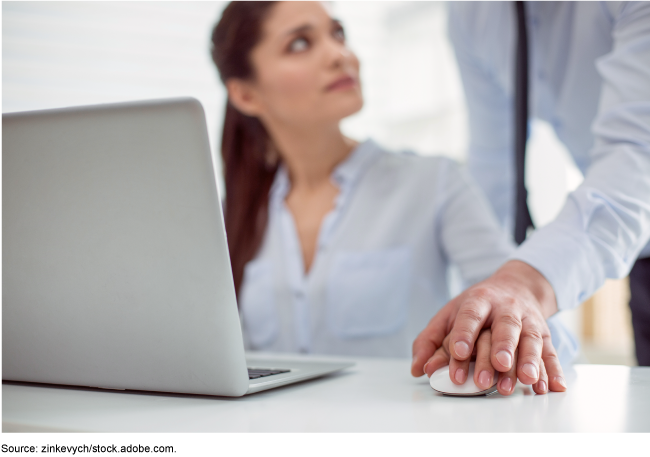 A male coworker leaning over a female coworker who is seated in front of a laptop