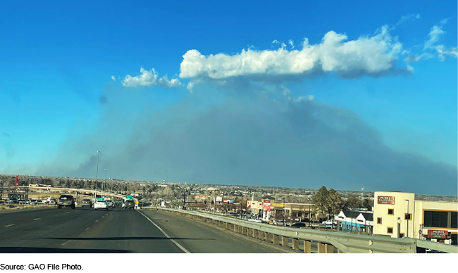 view from a highway with buildings on the right-hand side of it and smoke covering a blue sky