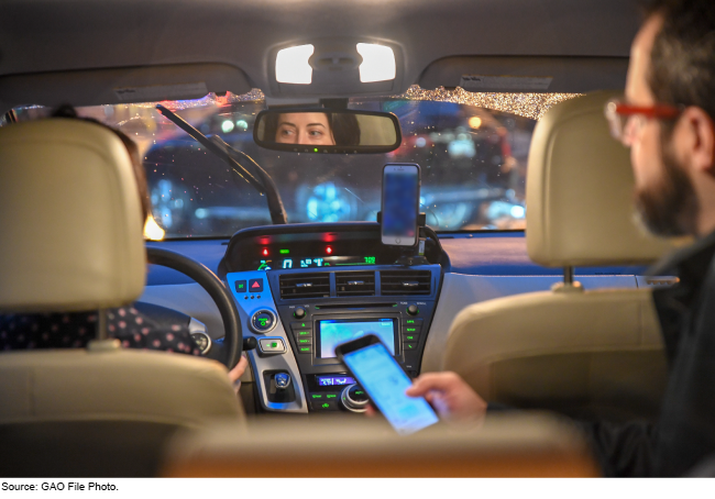 The inside of a car with a driver and a person in the back seat holding a cellphone.