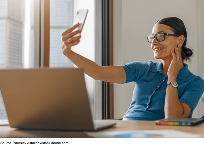 A woman sitting in front of a computer using a cell phone to take a photo of herself.