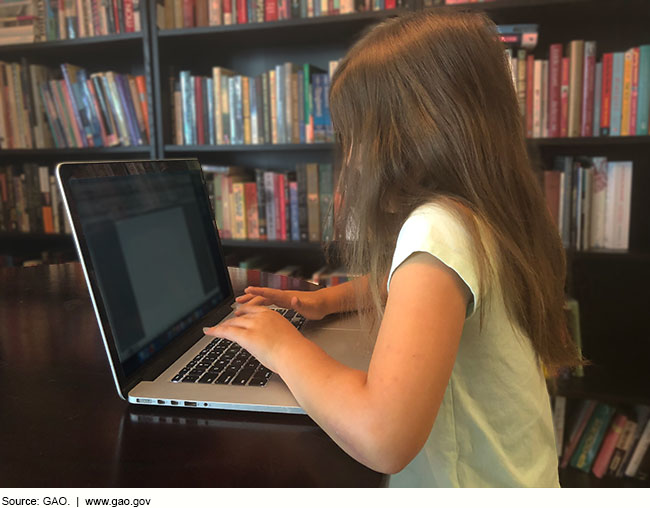 A child types on a laptop in front of a bookcase filled with books