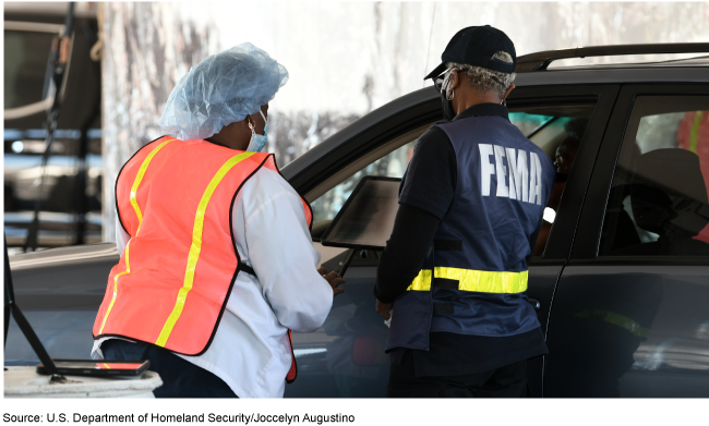 FEMA worker and a person in medical attire talking to a driver in a car.