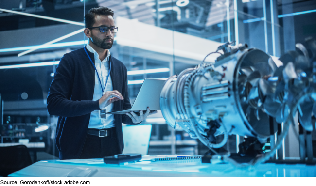 A scientist standing with a laptop open in front of a piece of equipment.