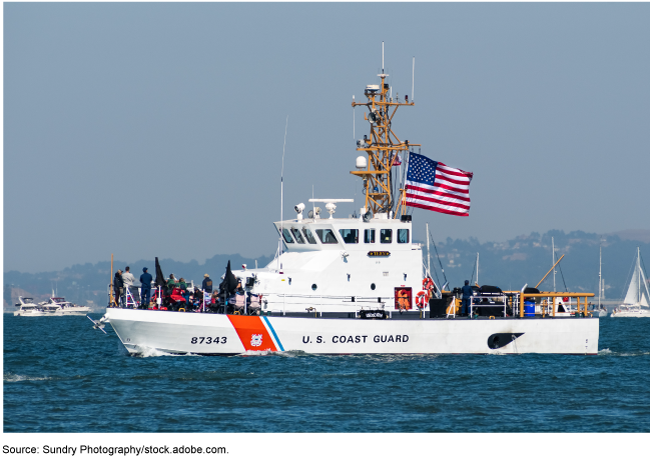 A U.S. Coast Guard cutter with people standing at the helm and an American flag waving in the wind.