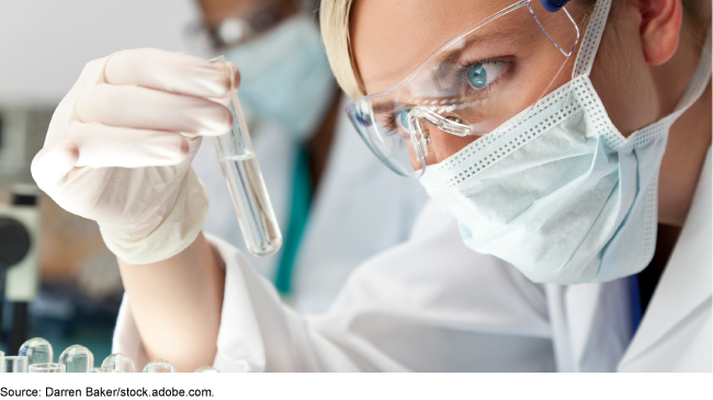 person wearing a lab coat, mask, gloves, and eye protection examines a liquid in a test tube