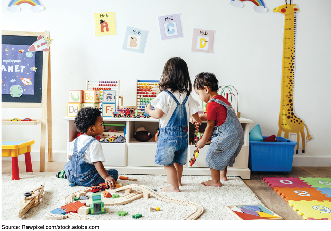 3 toddlers on a carpet in a room surrounded by toys, letters, and blocks.