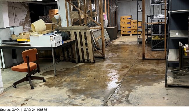 water on a cement floor of a room with a table, office chair, shelving, fallen lamp, boxes, wooden pallets, etc.