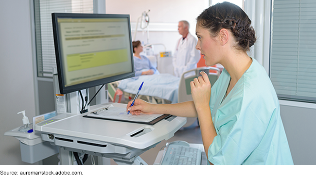 A person in medical scrubs taking notes and typing information into a computer near a hospital room.