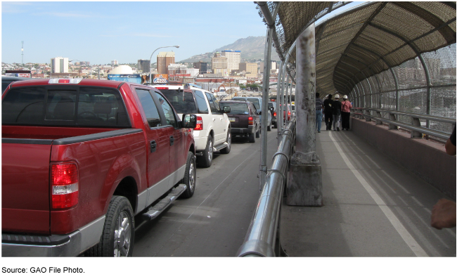 Photo showing a line of vehicles  stopped on a roadway with a walkway for people on the right.