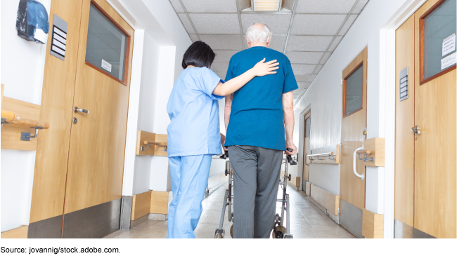 Elderly patient using a walker and a medical staff member walking down a hallway.