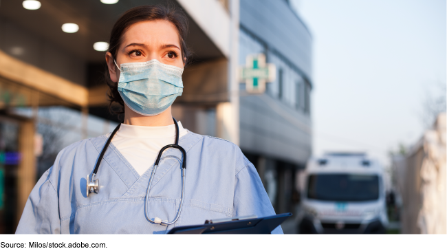 A medical person wearing scrubs and a mask stands outside holding a clipboard.