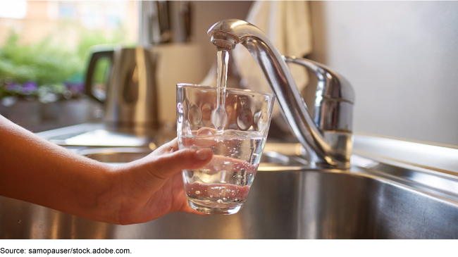 Close up of a person's hand and arm filling a glass with water under a sink faucet.