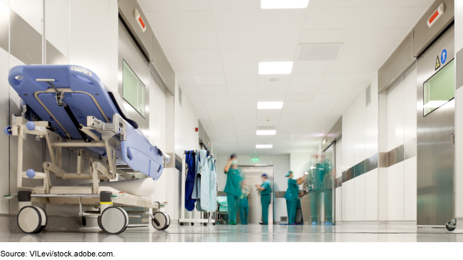 Photo showing a hospital hallway. In the foreground there's an empty gurney. In the background there are hospital workers in surgical scrubs.