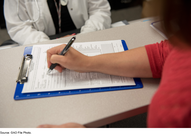 Photo showing a health care worker sitting across from a patient who is filling out forms on a clipboard.