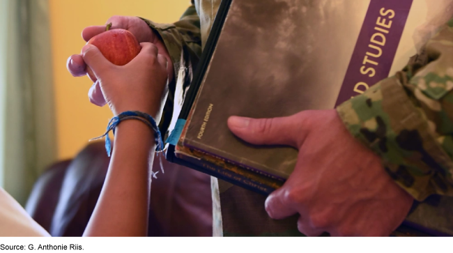 A child giving an apple to a person in a military uniform holding a textbook