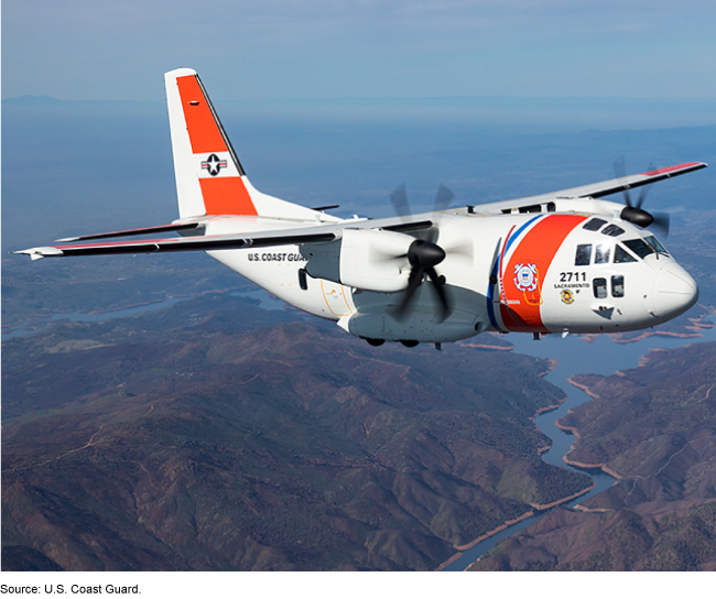 U.S. Coast Guard aircraft flies over hilly coastline.