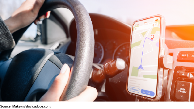 Hands on a car steering wheel with a mobile phone showing GPS navigation mounted to the dashboard
