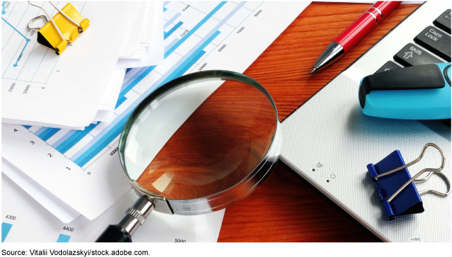 Magnifying glass resting on a keyboard with financial papers scattered around and below it.