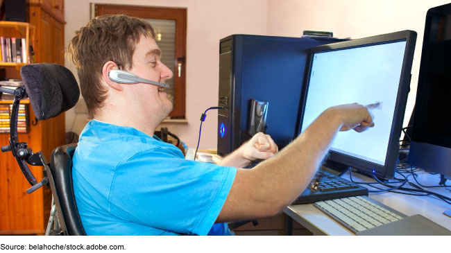 A young man wearing a headset sits at a computer and points to the screen