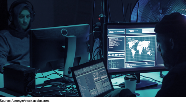 Stock image showing two people sitting in a dark room that is lit by their computer screens who seem to be hacking into a network. 