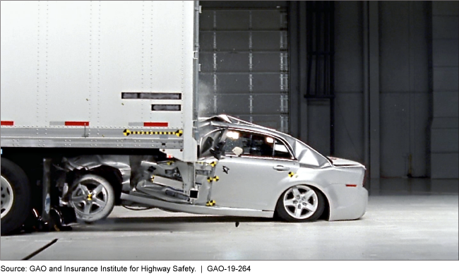 The passenger compartment of a silver test sedan is crushed beneath the back of a tractor-trailer in a simulated crash.