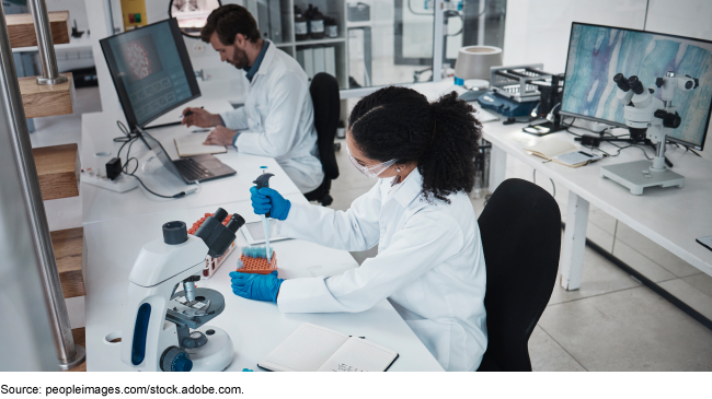 An image of two medical professionals sitting in a lab. One is working at the computer, examining notes. The other is preparing samples to be viewed under a microscope. 