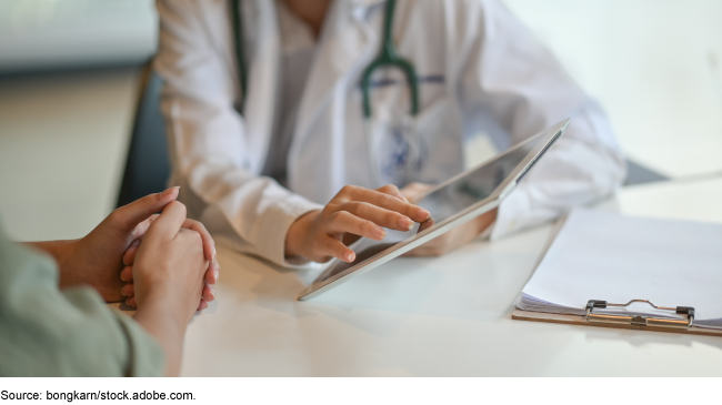 A person wearing medical jacket sitting across from someone while they both are looking at a tablet.