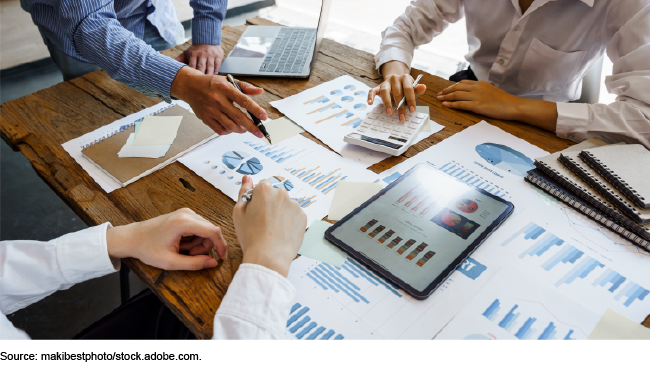 A photo showing 3 people's hands over a conference table covered in paperwork with charts and graphs