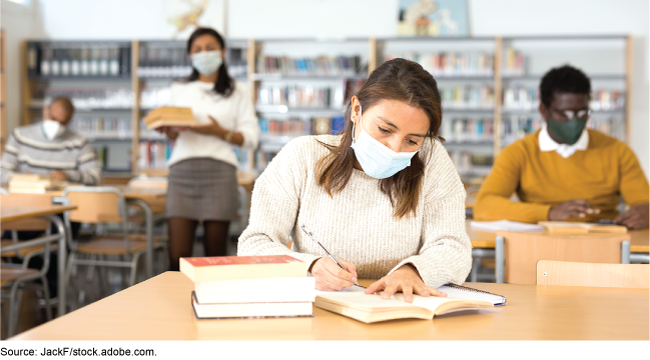 A woman wearing a mask works at a desk in a library with others in the background.
