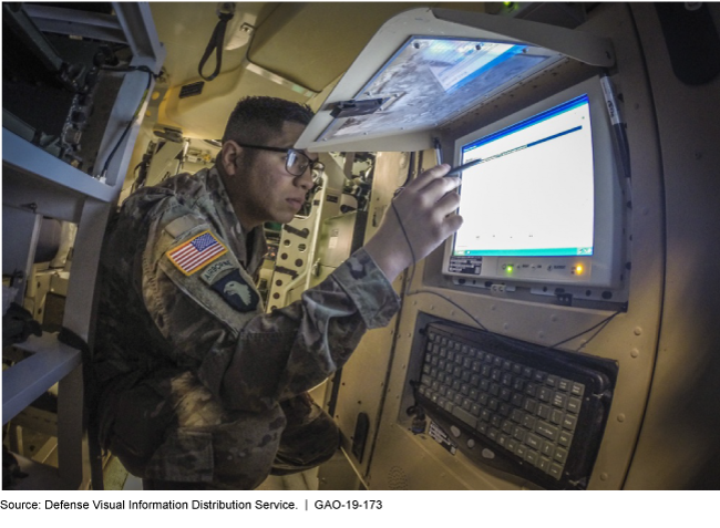 Photograph of a man in a military uniform using a computer that is mounted on the wall of a small space.