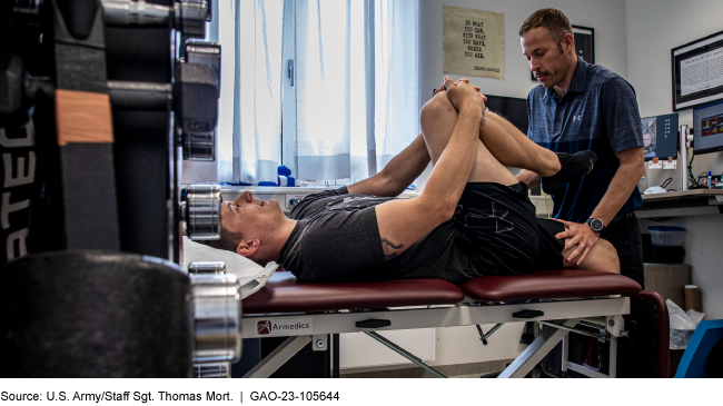 A man laying on his back on a medical exam table with his right leg bent at the knee and pulled across his left leg while a physical therapist looks on