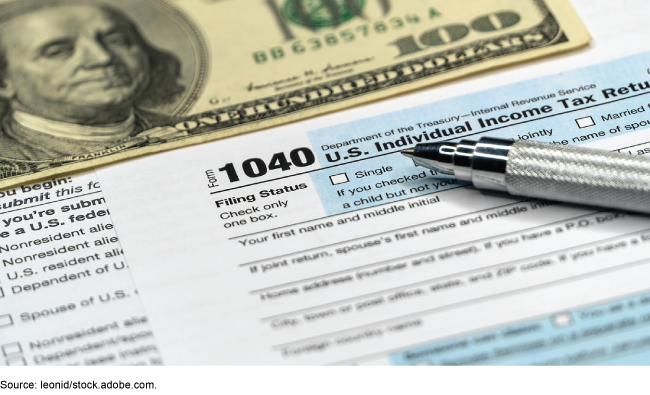A photograph with a one hundred dollar bill on top of an IRS tax return form and a pen.