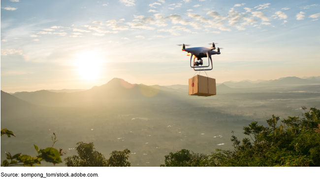 A drone carrying a package flies over trees with mountains in the background.