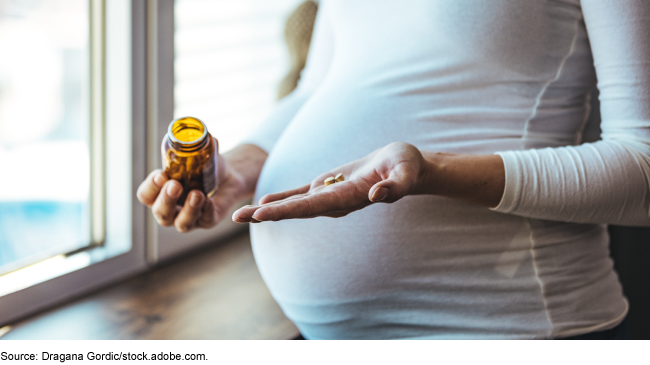 Close up of a pregnant woman's abdomen. She's holding a pill bottle in one hand and two pills in the other.
