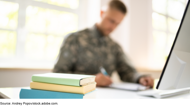 A person dressed in military fatigues sitting at a desk with a laptop, books, and clipboard in front of him.