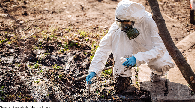 A scientist dressed in protective gear collecting a sample from the ground.