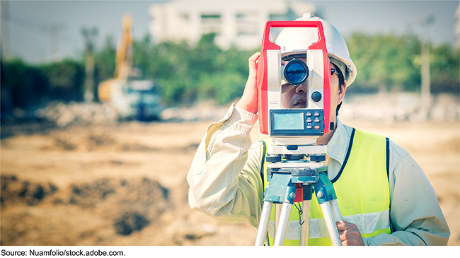 Front view of a person wearing a hardhat looking into a monocular on a surveying tripod at a work site. 