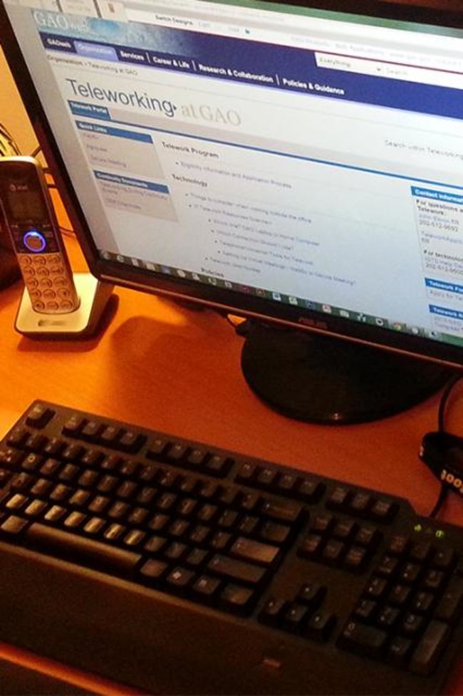 A home office desk with a telephone, keyboard, and computer monitor showing a webpage about GAO telework