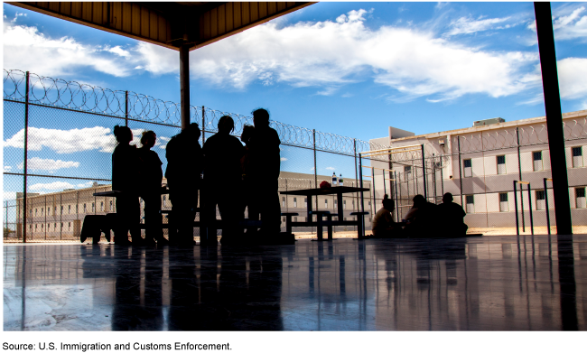 Silhouettes of people standing and sitting in a cement courtyard near a fence with razor wire on top