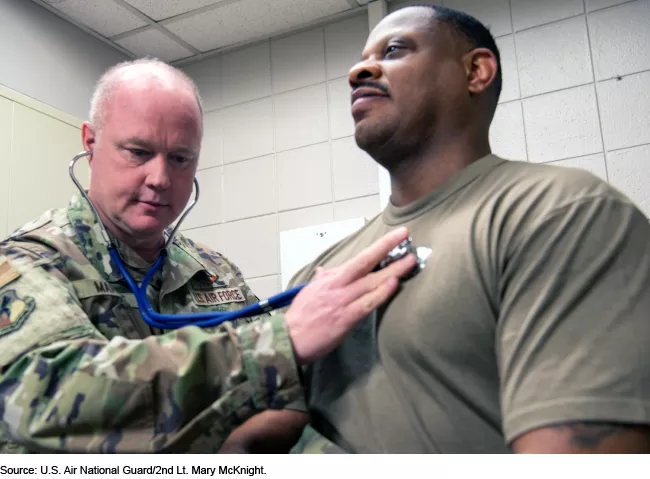 Photo showing a military doctor in fatigues using a stethoscope to listen to a service member's heart.