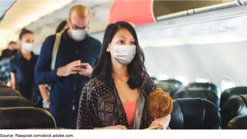 A woman wearing a face mask walks down the aisle of a plane, followed by several other people who are also wearing masks.