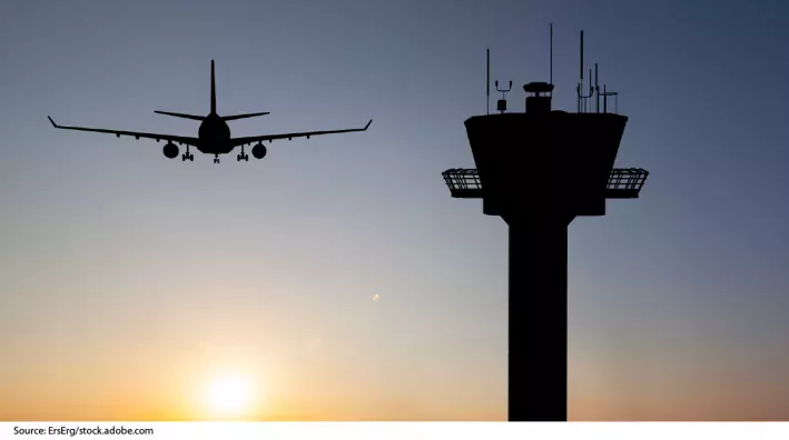 Photo showing a plane flying in the distance and an air traffic control tower in the forground.