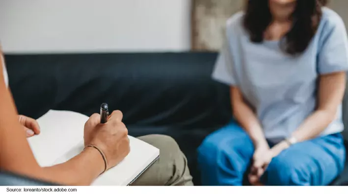 Stock image showing two people sitting across from each other. Nearest to us we see the arm and lap of a person with a clipboard taking notes. In the background, we see the neck-down of a person seated with their hands clasped.