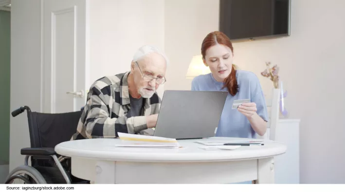 Photo showing an older adult sitting in a wheel chair next to a younger person. They are both working on a laptop together. 