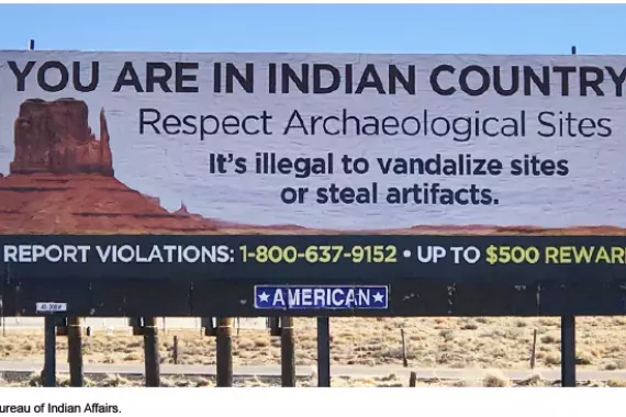 A billboard in a desert environment reads "You are in Indian Country. Respect Archaeological Sites. It's illegal to vandalize or steal artifacts. Report Violations, Up to $500 Reward.