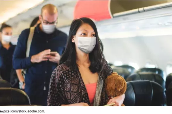 A woman wearing a face mask walks down the aisle of a plane, followed by several other people who are also wearing masks.