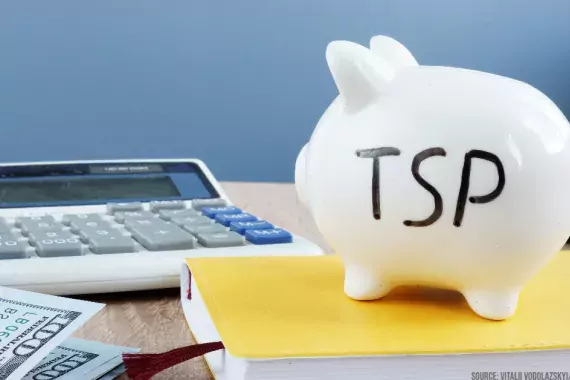 Photo showing a piggy bank with "TSP' written on it in the foreground and a calculator in the background