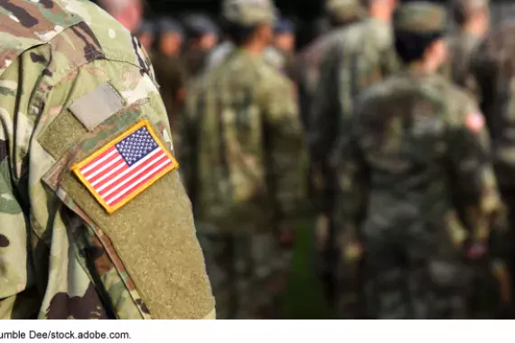 Photo showing the shoulder of a military servicemember in fatigues with a U.S.A. flag patch on arm. Other servicemembers are in the background.